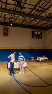 Two men are standing in a middle school gymnasium basketball court. They are standing in front of a table with their chests puffed up. They look as if they are about to fight.