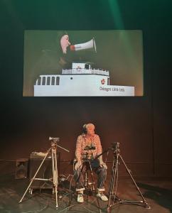 A woman sits below a screen surrounded by 3 video cameras. Above her on the screen is person wearing black with a white boat called Chicagos little lady built around their waist. They are yelling into a megaphone.