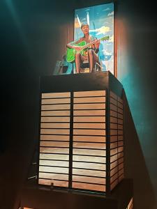 A person sits atop a tall box structure representing the Sears Tower. The person is also a part of the Sears tower wearing it's spires on top of their head.  They are holding a bright green guitar and are singing.