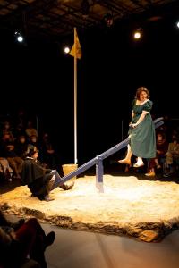 A girl in a dress is suspended at the top of a see-saw, there is a man sitting on the other end in the sand.