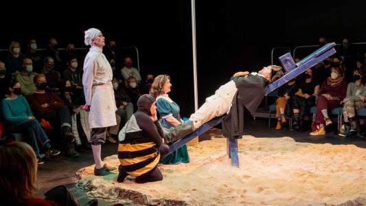 Napoleon lays on a seesaw with his head tilting up. Gathered near his feet are a chef, a girl and a person dressed as a bee. 