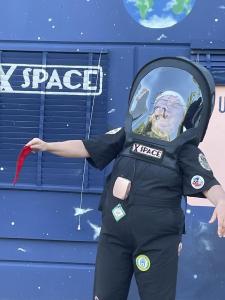 A space person wearing a huge black space helmet is laughing and holding a tiny red flag in their right hand.