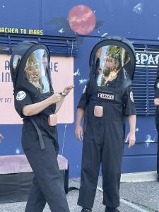 Two space people wearing huge black space helmets are standing in front of a wall that is painted to look like outer space.  One of them is holding a white marker and has been drawing all over the other persons space helmet over over their face.