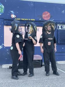 Three space people wearing huge black space helmets are standing in front of a wall that has been painted to look like outer space. Above them on the wall there is a tiny rocket that is on top of a track that says “Flight tracker to Mars”.