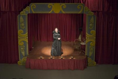 A woman in 1800s black funeral dress stands in a spotlight on a wooden stage framed by a yellow and grey decorated proscenium. The stage has four footlights and red curtains.
