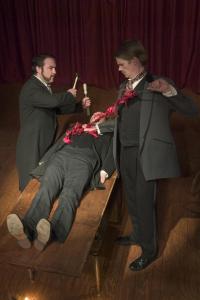 A man is lying on his back on a wooden table with a man standing on either side of him. They are all dressed in 1800s black and grey suits. The dark-haired man on the left is holding a hammer and chisel as if about to strike the chest of the man lying on the table. The other man is pulling out a long strand of red fabric pieces tied together from the abdomen of the man on the table.