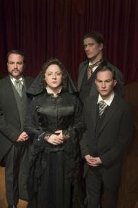 A woman and three men in 1800s funeral attire are looking forward in front of a red curtain.