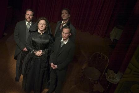 A down shot of a woman and three men all dressed in black and grey 1800s funeral attire are looking up and out seriously while standing on a wooden floor. Behind is a red curtain. To the right of them, in the shadows, is small side table with a glass and gold tray next to a wooden chair.