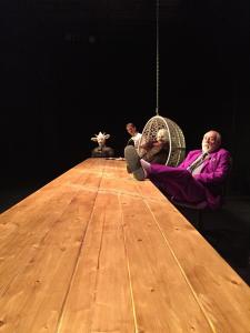 The camera sits at one end of a very long wooden table. On the other end is an odd foursome. Closest is an older man in a magenta suit who has his feet up on the table. Next is an eagle in a t-shirt sitting in a hanging wicker chair. Peaking from behind the chair is a man in a white shirt. Next to him is a cow in a dress.
