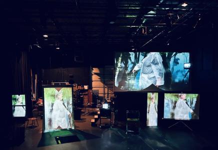 A behind the scenes look at the set- A far away photo of the warehouse and set which includes 5 surfaces with projections, stairs up to a platform, a floating tv, a patch of astro turf, and an A/V cart. 