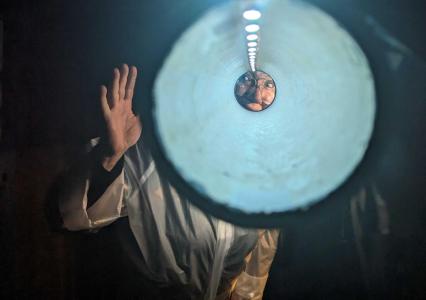 Looking through a tube with lights lining the top of it, at the end is the eyes and nose of a person. outside of the tube you can see the person waving. 