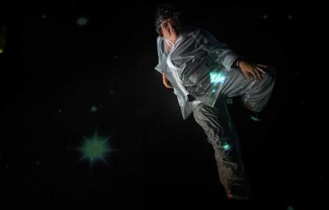 A down angle photo of a person in a milky clear plastic suit laying in blackness with stars projected on and around them.