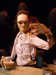 A life-size puppet of Tommy Lee Jones sits at a table in a diner.  Behind him, you can faintly see the silhouettes of 3 puppeteers dressed in all black who are operating the puppet.  Tommy Lee Jones is holding up a gold pocket watch which is on a gold chain. The puppet is looking at the watch.