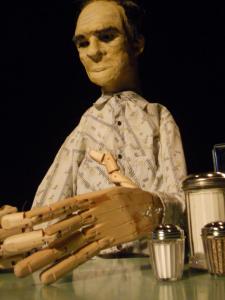 Close up of a life-size puppet of Tommy Lee Jones sitting at a diner table. His hands are wooden and are in the foreground.  They are built to work the same way as human hands and you can see the mechanism is uses fishing line and rubber bands.