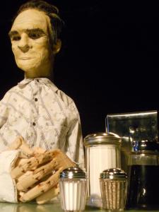 Close up of a life-size puppet of Tommy Lee Jones sitting at a diner table.  He looks out into the distance holding his wooden hands in front of him. One of his arms is in a cast, like his hand is broken.