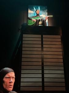 A tall box representing the sears tower has a person on top wearing the spires that are the top of the sears tower. They are holding a green guitar and singing. To the lower left of the tower a persons head pokes up with a confused face.