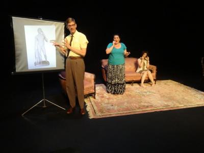 A man from the 1950's stands at a projector screen gesturing to an image of a statue of Herakles. Behind him, a woman from today stands on a pink carpet talking on a cell phone. Behind her, a woman from the 1920's sit on a couch looking into a cosmetic mirror.