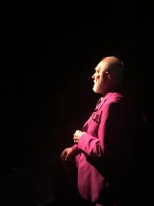 Surrounded by darkness, an older man in a magenta suit is speaking in profile. He is starkly lit from the front, creating a chiaroscuro effect.