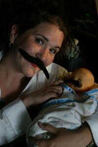 A woman with a fake mustache cradles a baby doll that also has a fake mustache.