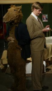A man in a brown suit looks at small paper scraps in his hand.  He stands back-to-back with a person in a camel costume. In the background are two beds suspended above the floor with wire.