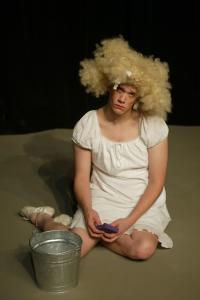A very tall and homely Cinderella sits on the floor looking forlorn with a huge blond curly wig and a mop bucket and sponge.