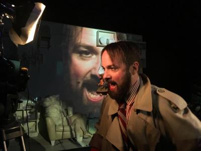 A bearded man in a trench coat is in profile talking passionately into a camera. Behind him, is a projection of the front of his face.