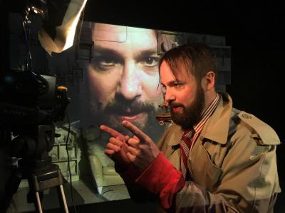 A bearded man in a trench coat is in profile talking into a camera. He has both of his pointer fingers up. Behind him, is a projection of the front of his face.