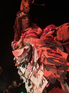 Upshot of a person covered in magazine pages. She is twisting in order to read from one of them. She has a large headpiece on that includes a plastic horse head with antlers. The lighting is starkly white and red.
