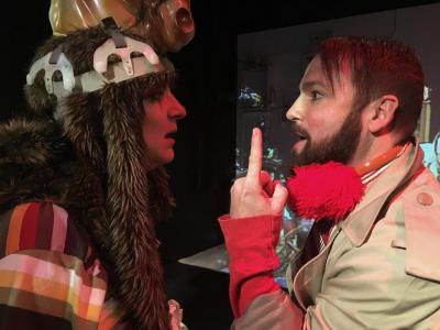 A close-up of two people arguing in profile. One wears a strange headpiece made of fake fur and a plastic horse head. The other wears a trench coat and has bright red earmuffs around his neck. He is holding his finger up.