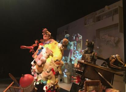 A person wearing a giant outfit made of stuffed animals stands in the middle of a cluttered room. She has her arms out and is talking. In the background there is a white wall full of white items.