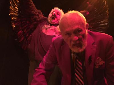 In stark magenta light, an older bearded man in a suit leans towards the camera, yelling. Behind him, a person dressed as an eagle with its wings spread is also yelling. The eagle is wearing a t-shirt.
