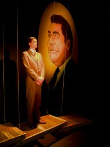 A man in a brown suit stands holding his hand together at his waist. He is looking up and to his left solemnly. He is standing on a narrow metal grated walkway that is suspended above the floor by wires. Behind is a mural of a man with black hair.