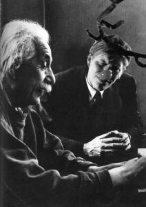 A man, with antlers and a red deer nose, sits with Einstein pondering space and time.