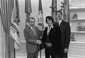 Elvis Presley stands in the oval office shaking President Nixon’s hand. Behind Elvis is a man with antlers and a red deer nose.