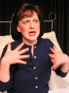 Close up of a woman sitting on a large bed wearing a blue button up sweater. Her long hair is gathered up in a hairnet 1940s style. She’s in the midst of saying something emphatic while gesticulating with both of her hands.