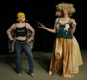 Cinderella, wearing a yellow skirt and a green top with clocks on the bust, is holding up a glass slipper to a woman wearing jeans, a mask, and a sparkly shirt that says "fairy".