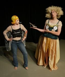 Cinderella, wearing a yellow skirt and a green top with clocks on the bust, is holding up a glass slipper to a woman wearing jeans, a mask, and a sparkly shirt that says "fairy".