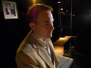 A blonde man in a tan suit with no tie stands in profile in the foreground. Behind him to the left is a photo of a man’s face with black hair hanging from wires to the ceiling. To the right of the man in the distance the is a narrow metal grated walkway extending into the dark in the distance. The man is looking concerned and is dramatically backlit by amber and white lights.