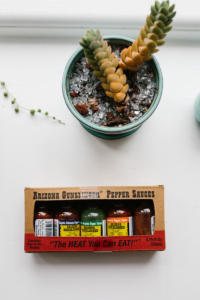 Down shot of a white surface with a potted succulent. Below the plant is a box of Arizona Gunslinger Pepper Sauces. 5 small bottles of hot sauce are in the box, which proclaims “The Heat You Can Eat!”Deep in the recesses of the refrigerator door lies the mythical Arizona Gunslinger! Sadly they expired 2 years ago before we ever even opened em. Wah wah.