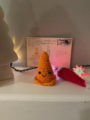 A postcard is displayed on a mantel. In front of the postcard is a knitted traffic cone with a smiling face stitched into it. What looks like a knitted Santa hat is next to the traffic cone.