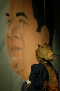 A person in a camel costume wearing a blue blazer looks forward while standing in front a large mural portrait of a man’s face with dark hair.