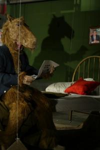 A person in a camel costume is seated on a bed with a red and white pillow on it. The bed is suspended by wires and the camel is reading a newspaper.