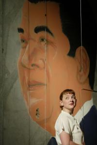 A woman with red hair and white shirt looks wistfully upward while standing in front of a large portrait of a man’s face with dark hair.