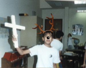 A boy, with antlers and deer nose, holds up a large white cross in a classroom.