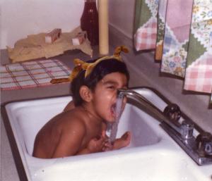 A young child, with antlers and deer nose, bathes in the kitchen sink, attempting to eat the water.