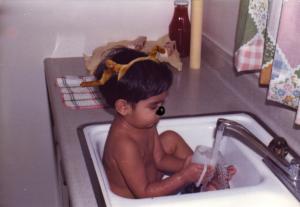 A young child, with antlers and deer nose, bathes in the kitchen sink. 