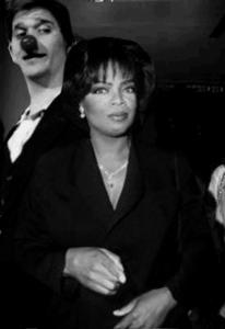 Oprah Winfrey stands center in the photograph. A man with a red deer nose stands behind her.