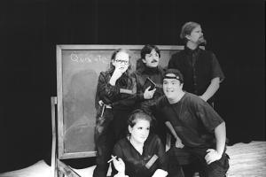 A ragged looking group of men in women dressed entirely in black, and covered in chalk dust, stand in front of a giant rolling chalkboard. On the upper left hand corner of the chalkboard, the word Quixote is written.