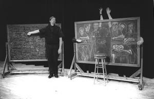 A professor with a large mustache and goatee stands in front of a chalkboard packed full of writing. Next to him is another chalkboard with a chalk drawing of a students in a classroom. In the middle of the chalk drawing there is a life size outline of a student who is missing. The name "Chip" is written in the blank spot.