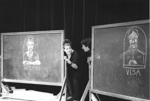 Two large, rolling chalkboards are pushed together. One board has a chalk drawing of a woman working at a passport window. The other board has a chalk drawing of a man working at a Visa window. A man and a woman peak out from the opening made between the two chalkboards. It is clear that the drawings on the chalkboards are modeled after them.